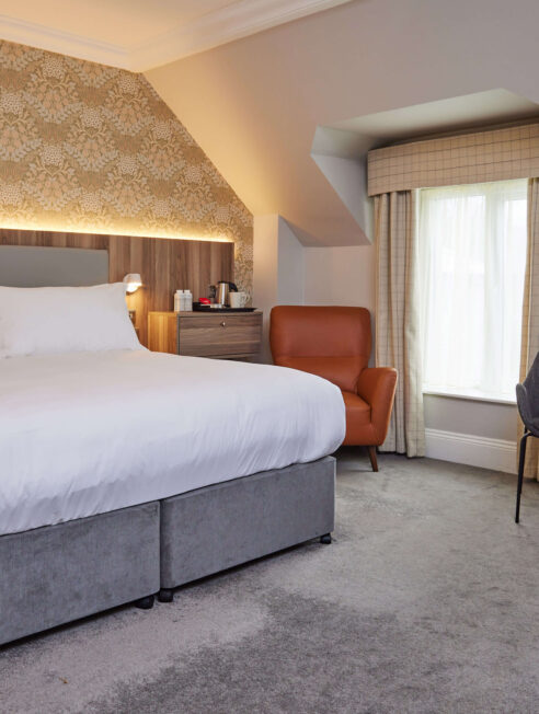 Spacious hotel room with king-size bed, plush armchair, elegant decor, and natural light, perfect for relaxation.