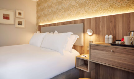 Cosy hotel room with plush bed, elegant wallpaper, warm lighting, bedside table with water bottles, and a coffee station.