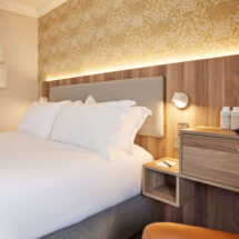 Cosy hotel room with plush bedding, warm lighting, stylish wallpaper, and amenities like bottled water and tea set.