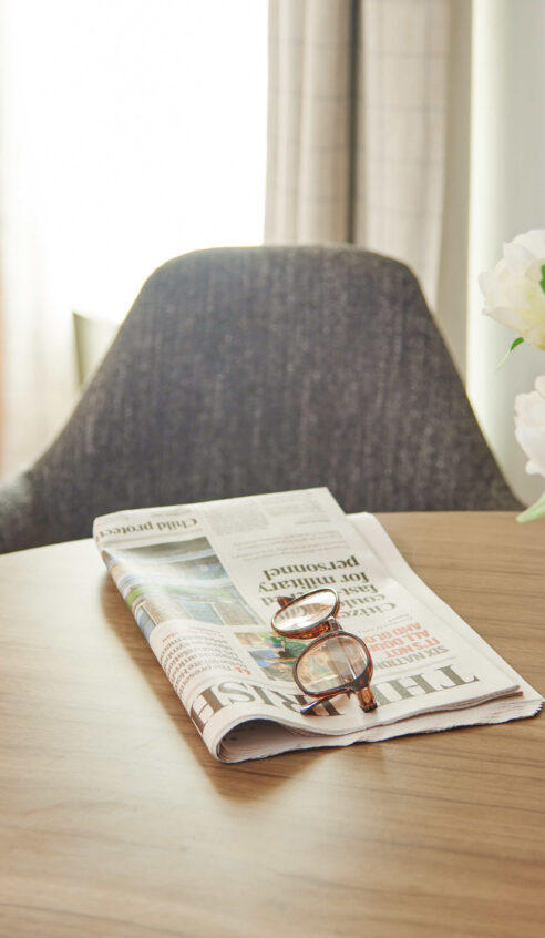Cosy hotel room with a stylish armchair, round wooden table, fresh flowers, newspaper, and ample natural light.