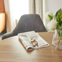 Cosy hotel room corner with an armchair, fresh flowers on a wooden table, and a newspaper welcoming guests.