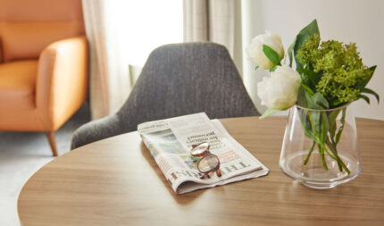 Cosy hotel room corner with an armchair, fresh flowers on a wooden table, and a newspaper welcoming guests.