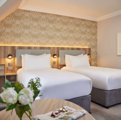 Twin beds in a modern hotel room with floral wallpaper, elegant lighting, tea amenities, white bedding, cosy atmosphere.