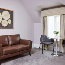 Cosy hotel sitting area with modern leather sofa, art decor, round table, two chairs, and natural light for relaxing stay.