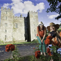 Guests admire vibrant roses in front of a historic castle under a clear blue sky, capturing memories on camera.