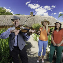 Guests enjoy live traditional music in vibrant garden setting beside charming thatched cottage with clear blue skies.