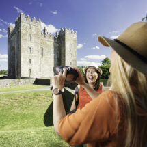 Guests smiling and taking photos at historic stone castle under clear blue skies, surrounded by lush greenery.