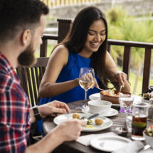 Guests dining on a sunny terrace with river views, enjoying gourmet dishes and wine at a luxury countryside hotel.