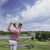 Guest enjoying a scenic golf course with lake and castle views, lush trees, clear skies, at a luxury countryside hotel.
