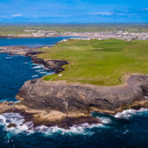 Breathtaking seaside view of rugged cliffs, lush green landscape, and a quaint coastal village under a clear blue sky.