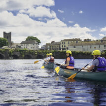 Guests canoeing on a scenic river with a historic stone bridge and charming town in the background, sunny blue skies.