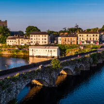 Scenic riverside village view with stone bridge, charming cottages, lush greenery, and a historic church in golden sunlight.