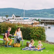 Family enjoys picnic by scenic lake with boats, lush greenery, and distant hills at welcoming hotel. Relaxing getaway spot.