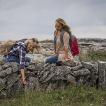 Two women explore a scenic countryside, kneeling on a stone wall with lush greenery, under a cloudy sky, enjoying nature.