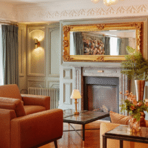 Elegant hotel lounge with cosy orange seating, ornate mirror, stylish lighting, and inviting fireplace. Relaxing atmosphere.