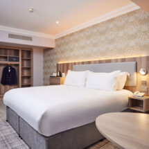 Elegant hotel room with plush king-size bed, soft lighting, desk chair, wardrobe, floral wallpaper, and fresh flowers.