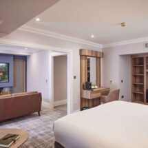 Spacious hotel suite with plush bed, cosy seating, modern decor, and an electric fireplace for a relaxing stay.