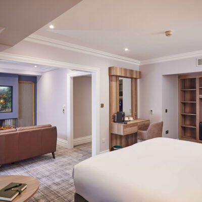 Spacious hotel suite with plush bed, cosy seating, modern decor, and an electric fireplace for a relaxing stay.