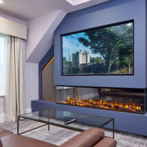Luxurious hotel suite with a cosy fireplace, plush leather sofa, large TV screen, and scenic window view.