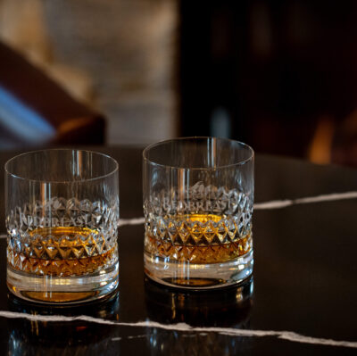 Two crystal glasses of whiskey on a polished marble table by a cozy fireplace in a luxurious hotel lounge.