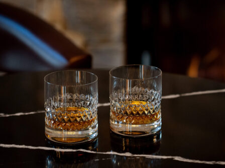 Two crystal glasses of whiskey on a polished marble table by a cozy fireplace in a luxurious hotel lounge.