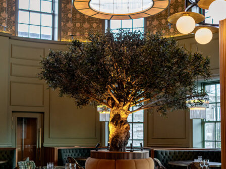 Elegant hotel lobby with central olive tree, warm lighting, plush seating, and stylish décor, offering a welcoming ambience.