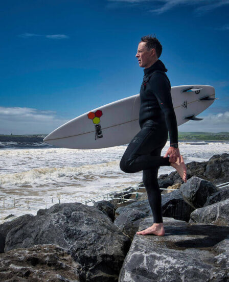 Surfer on rock overlooking ocean waves, surfboard in hand, under a clear blue sky, capturing coastal adventure near hotel.