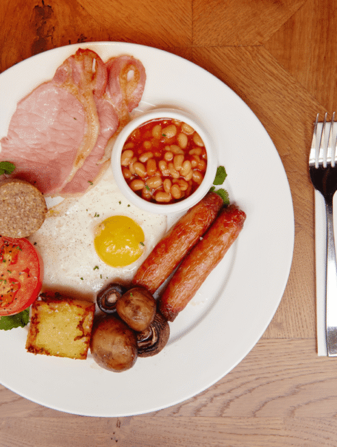 Traditional full English breakfast with bacon, sausages, egg, baked beans, and toast on a wooden dining table.