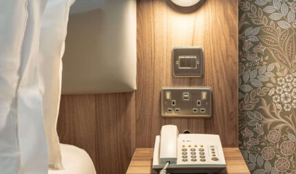 Elegant hotel bedroom with modern lighting, plush bedding, bedside table, and classic wallpaper, offering comfort and style.