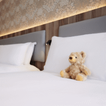 Luxurious double bed with plush teddy bear, elegant headboard, soft lighting, and patterned wall in cosy hotel room.