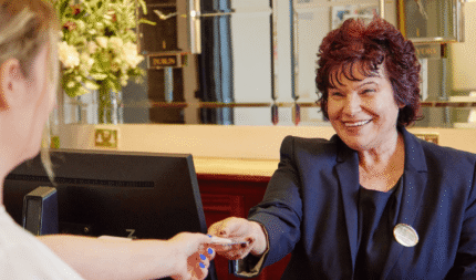 Friendly hotel receptionist offering keys at elegant wooden counter with lush floral arrangement in the lobby.