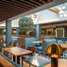 Elegant hotel dining area with cosy blue booths, warm wooden tables, ambient lighting, and a central decorative tree.